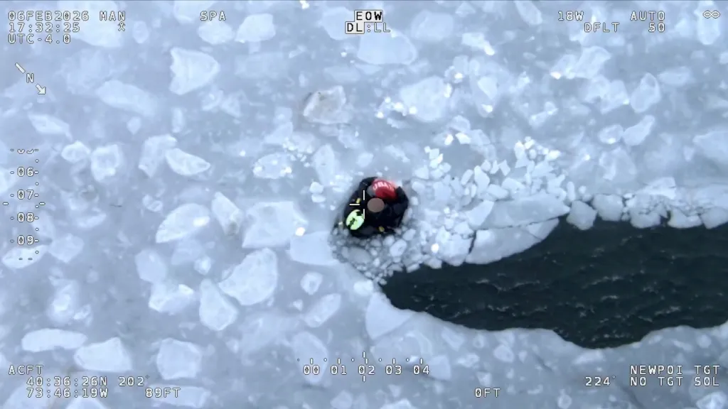 Aerial view of a person in a black suit with a red life vest in icy water, surrounded by ice floes.
